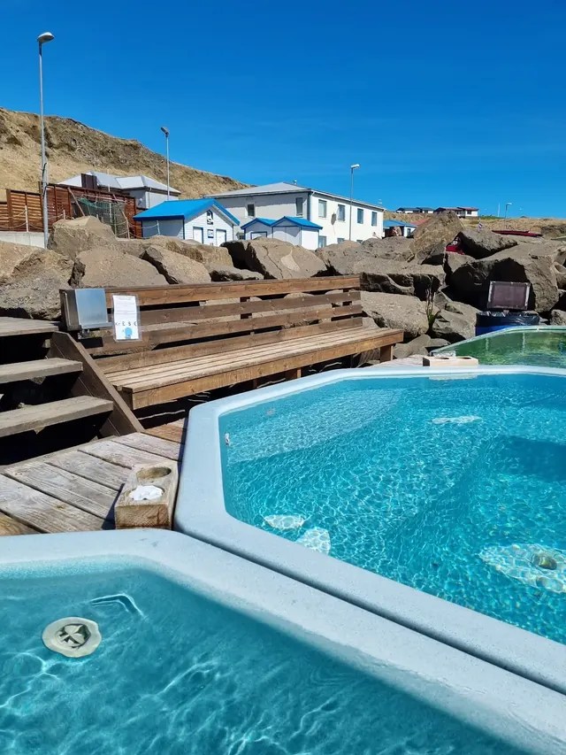 Clear blue water in octagonal hot spring pools with wooden benches and white buildings in the background at Drangsnes Hot Pots.