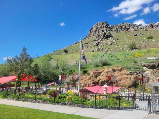 Daytime wide-angle view of Aura Soma Lava Hot Springs in Lava Hot Springs, Idaho, with vibrant red canopies, green landscape, and rocky mountain backdrop.