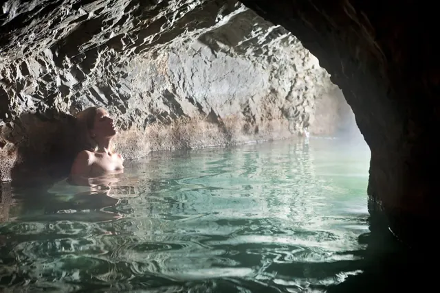 Person relaxing in a steamy indoor hot spring pool inside a rocky cave at QC Terme Bagni Vecchi in the Italian Alps showing textured stone.