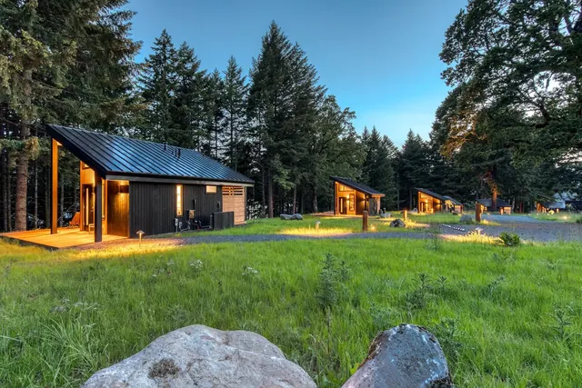Exterior view of multiple modern dark wood cabins with metal roofs lit at dusk surrounded by grassy field and tall trees at Tenzen Springs.