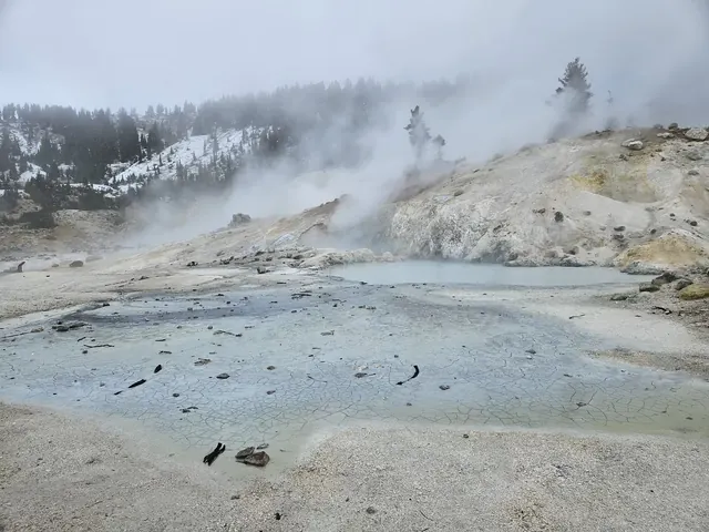 Foggy scene at Bumpass Hell Trail showing a cracked earth mineral pool with steam rising and snow dusting the nearby forested hills.