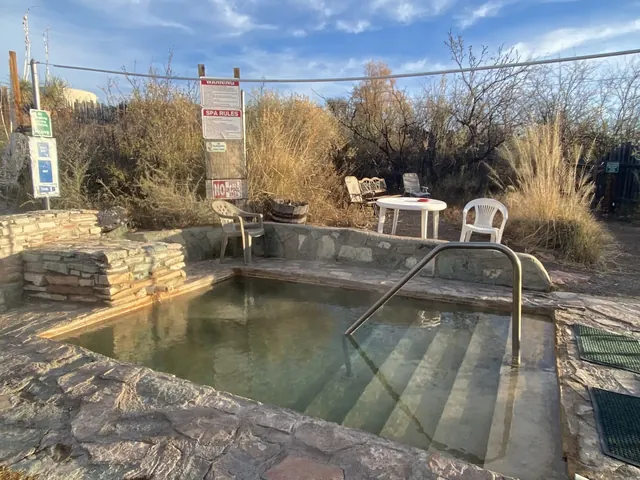 Small stone-bordered thermal pool at Faywood Hot Springs surrounded by dry brush and plastic chairs near Deming New Mexico.