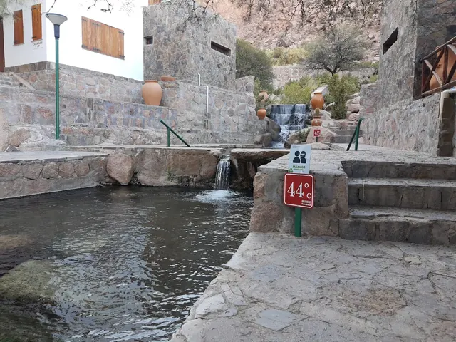 Stone pathways and multiple small hot spring pools with temperature signs and mountain cliffs visible at Termas de Fiambalá, Argentina.