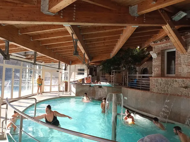 Indoor hot spring pool with wooden beam ceiling at Les Bains de Llo showing people enjoying the water and a wall of large windows letting i.
