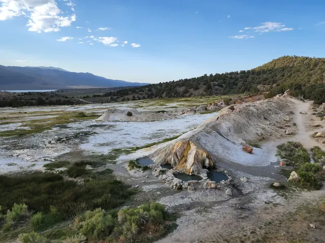 Wide view of Travertine hot springs showing multiple mineral pools surrounded by grassy and rocky terrain with mountains in the background.