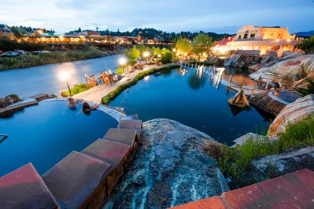 Evening view of an outdoor pool area with resort buildings lit up near river at The Springs Resort & Spa in Pagosa Springs, Colorado