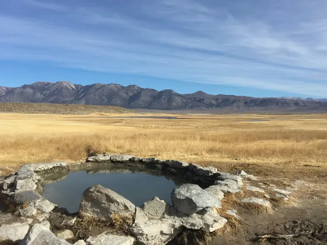 Crab Cooker Hot Springs natural stone pool filled with water in dry grassland with Mammoth Lakes mountains in California background.