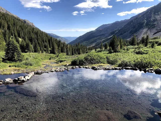 Clear water pool of Conundrum Hot Springs surrounded by lush greenery and pine trees near Aspen, Colorado, under a blue sky.
