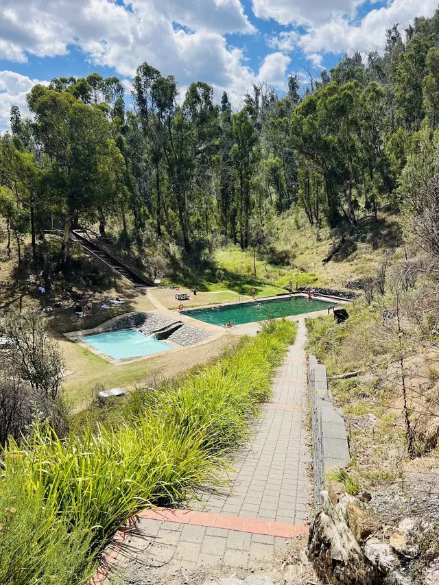 Stairs lead down to a green outdoor pool and a smaller thermal pool at Yarrangobilly Caves thermal pool, surrounded by grassy banks and tal.