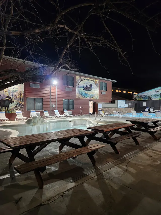 Night view of empty spa pools and long wooden picnic tables outside Spa Hot Springs Motel and Clinic with moose mural visible.
