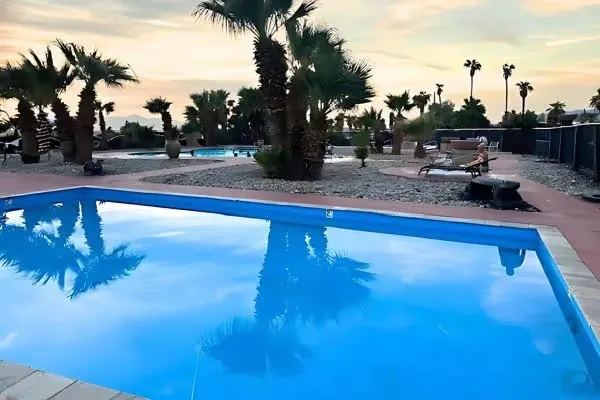 Calm reflecting pool in a hot spring area at dusk with tall palm trees, lounge chairs, and desert landscaping at Glamis North Hot Springs R.