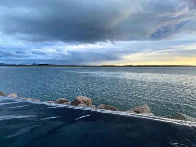 View of the dark water edge of the hot spring pool looking out across a calm large body of water with distant hills under moody clouds at S.
