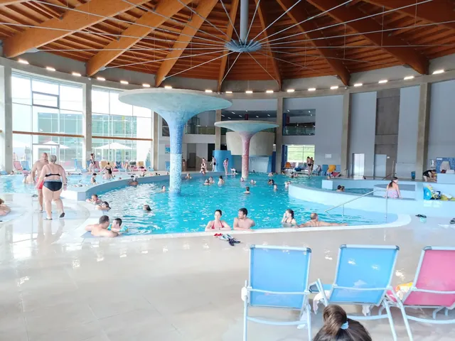Indoor hot spring pool at Termaly Losiny showing many people swimming under a wooden ceiling with large windows on one side.