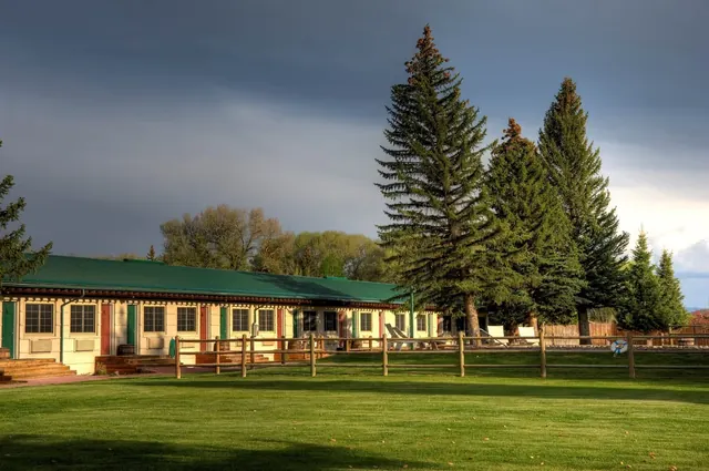 Long building with green roof and multiple doors facing a fenced grass area with tall pine trees at Saratoga Hot Springs Resort in Wyoming.