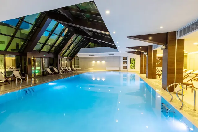 Large indoor thermal pool with blue water, glass roof, and rows of lounge chairs at Thermal Aqua Park Persenk in Devin, Bulgaria.