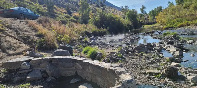 Stone-walled hot spring pool emitting steam next to a flowing river and green vegetation at Renova Hot Springs, Montana.