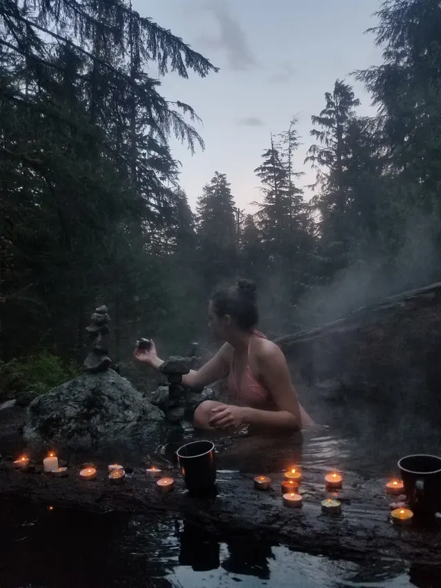 A woman sits quietly in a steaming hot spring pool at dusk with lit candles and stone stacks along a log edge under tall forest trees.