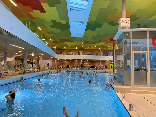 Indoor hot spring pool at Therme Wien in Vienna Austria with colorful ceiling panels and many people swimming and relaxing in the water.