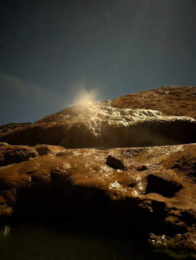 Steam rising from warm Red Hill Hot Springs mineral pools at night under a starry sky with light reflecting off rocky formations.