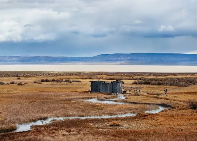 Rustic bathhouse by steaming hot spring on grassy prairie at Alvord Hot Springs, with distant mesa hills and cloudy sky in southeastern Ore.