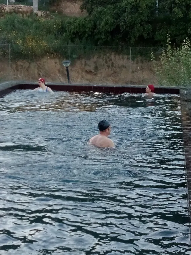 Three people wearing swim caps enjoy an outdoor mineral soaking pool surrounded by green trees at Termas de Longroiva.