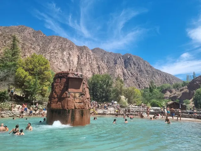 Large crowded hot spring pool at Termas de Cacheuta with a central stone water feature surrounded by rocky mountain landscape in Mendoza.