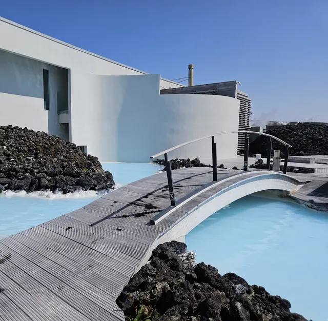 Wooden boardwalk and arched footbridge over bright blue water next to white building and black lava rocks at Blue Lagoon Iceland.