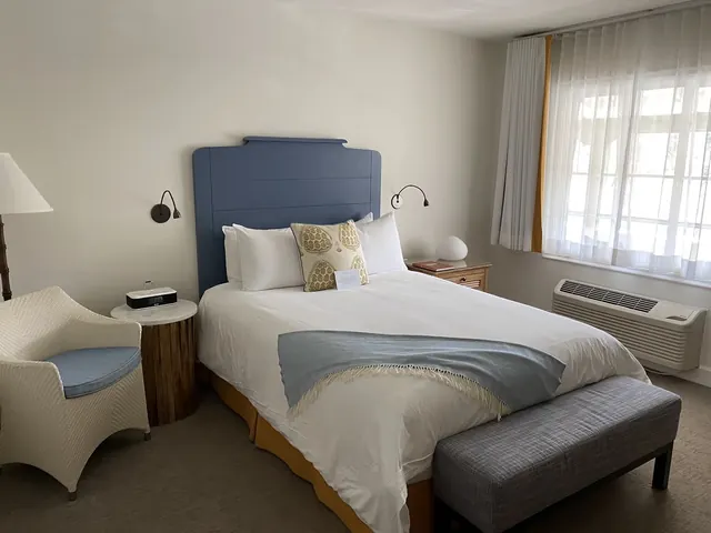 Bright hotel bedroom with white walls, a blue headboard, a cozy chair, and a large window with curtains letting in natural light.