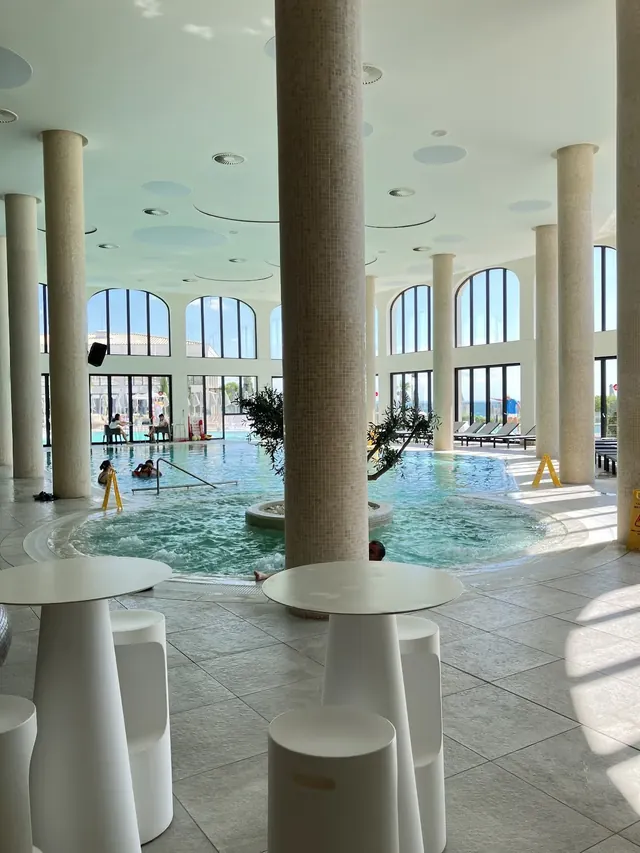 Indoor spa pool at Aquahouse Thermal and Beach with arched windows, tiled columns, steaming water, and white tables and stools nearby in Va.