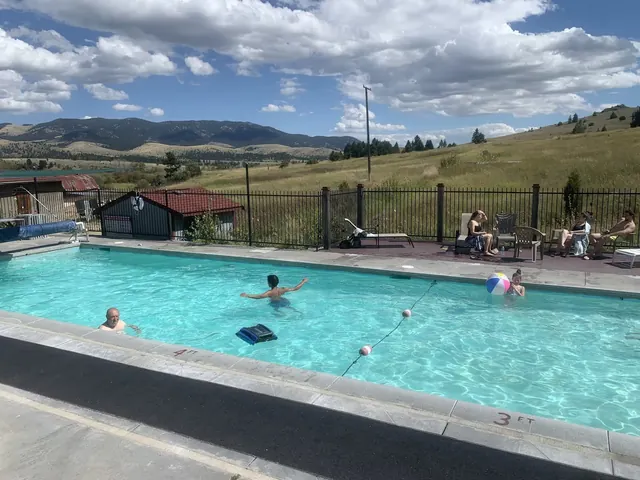 Boulder Hot Springs outdoor pool with several people swimming and sitting under partly cloudy skies with mountain views outside Boulder Mon.