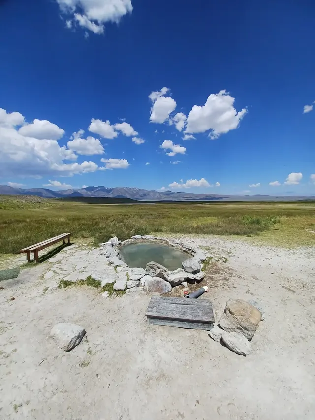 Round stone hot spring pool with water and a wooden bench in open grassy plain under blue sky at Shepherd Hot Springs.
