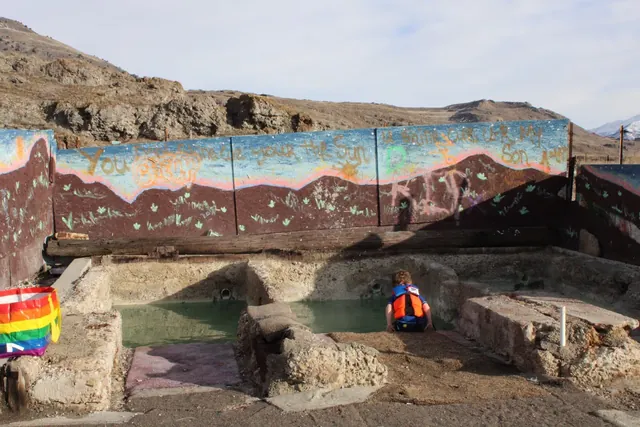 A child in a life jacket leans over a shallow pool at Old Indian Hot Spring, with colorful painted walls and rocky hills in the background.
