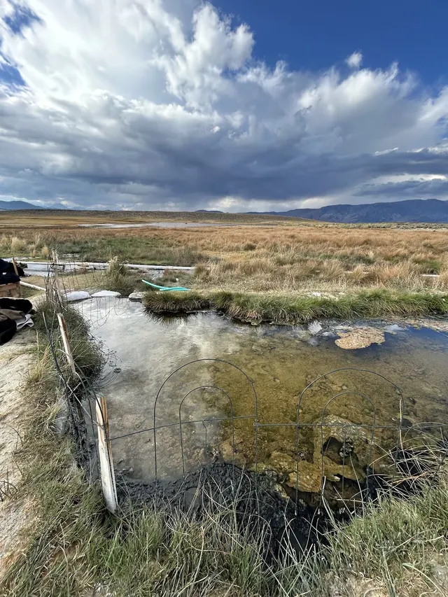 Natural hot spring pool surrounded by grass with a wire fence and a cloudy sky at Hilltop Pulky's Pool near Mammoth Lakes California.