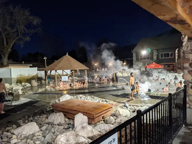 Night view of Crystal Hot Springs with illuminated pools, steam rising, people relaxing, and a wooden pavilion under dark sky.