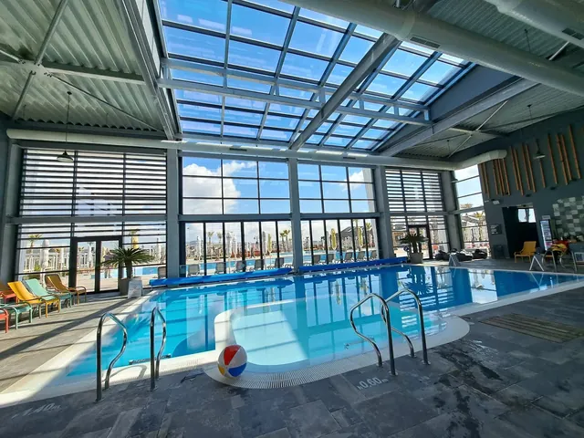 Indoor pool at Terme di Hissar in Hisarya Bulgaria with clear blue water, large glass windows, metal ceiling beams, and colorful chairs nea.