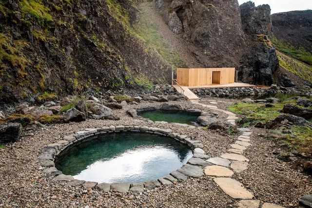 Clear water outdoor stone-edged soaking pools at Husafell Canyon Baths with rocky terrain and wooden changing hut nearby.