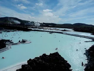 Large Blue Lagoon outdoor mineral pool with distant people soaking and steam rising against Icelandic hills near Grindavik.