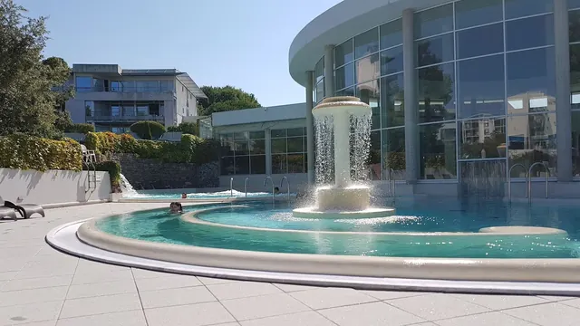 Outdoor spa pool at Sourceo France with round fountain spraying water under bright sunlight near large glass building.