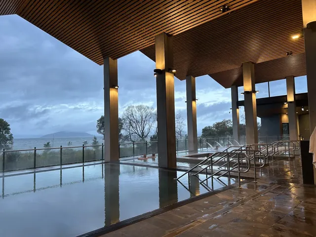 Multiple steaming hot spring soaking pools at Wai Ariki with metal handrails and glass fencing facing a lake and cloudy sky in Rotorua New.