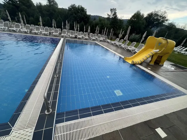 Outdoor kids pool at Regnum Banya Thermal Pools featuring a yellow elephant slide and multiple white lounge chairs with forested hills in t.