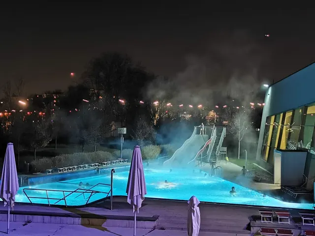 Nighttime view of an outdoor hot spring pool at Therme Wien in Vienna Austria with steam rising and a water slide near lounge chairs.