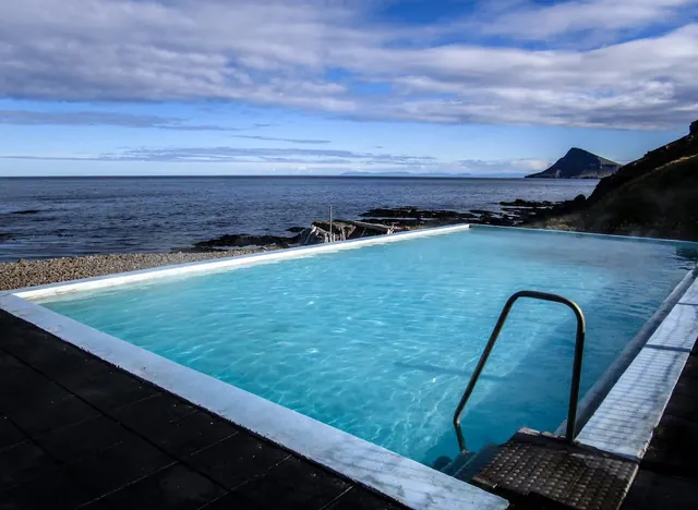 Swimmers enjoying the blue water of Krossnes Swimming Pool with a wooden fence and rough coastline in the background near Nordurfjordur, Ic.