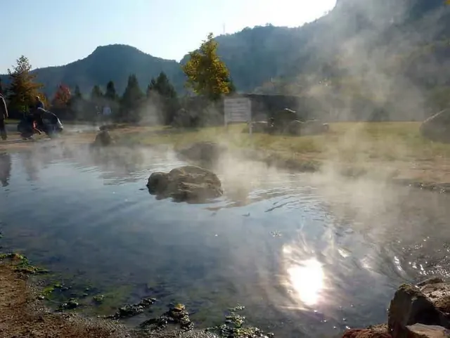 Steam rising from a large outdoor hot spring pool with mountainous landscape and scattered trees in the background at Rupite Thermal Spring.