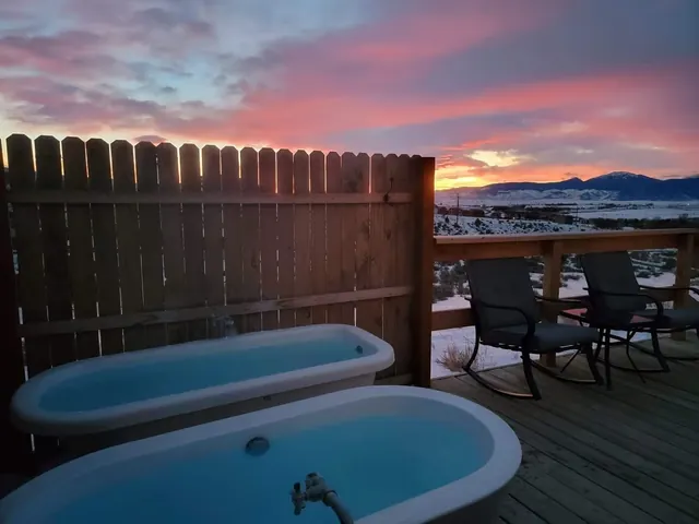 Pair of clawfoot tubs on a wooden deck with chairs and a fenced area overlooking a snowy mountain range at Pipestone Hot Springs in Montana.
