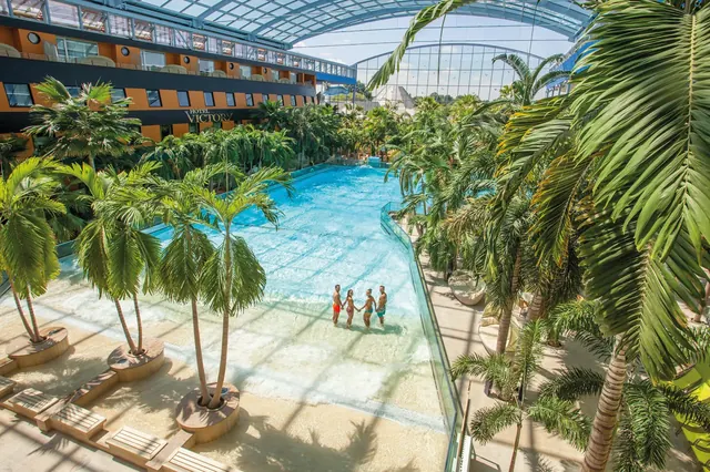 Indoor hot spring pool at Therme Erding, Germany, surrounded by lush palm trees and a hotel building under a large glass roof.