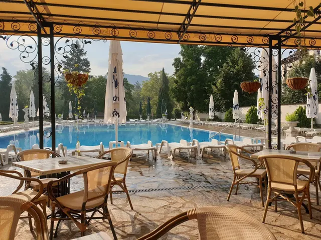 Covered patio seating area with wooden tables and chairs looking out to circular hot spring pool and umbrellas in Bulgaria.