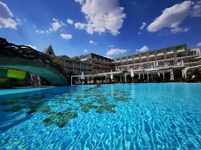 Clear blue water in an outdoor hot spring pool at Therma ECO Village Mineral Pools with a curved wooden bridge and hotel balconies in Varna.